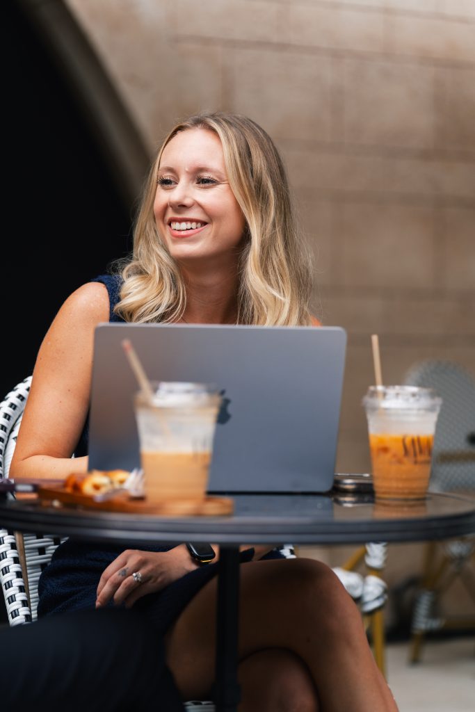 Isabel Allen of Team Sigh smiling at an outdoor café table with a laptop and iced coffee